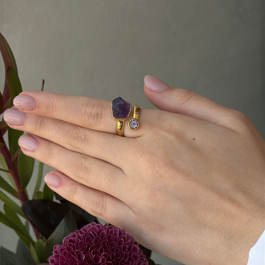 Hand wearing a gold ring with a purple gemstone, holding a purple flower against a neutral background